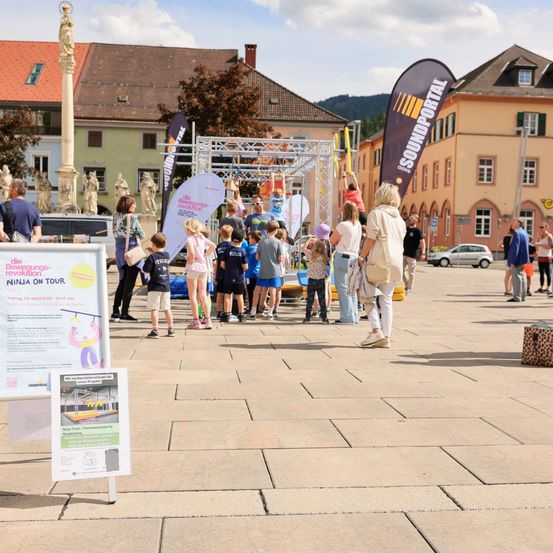 Eine Gruppe von Menschen steht in einer Plaza in der Nähe einer Bühne mit mehreren Bannern. Kinder spielen auf der Bühne mit verschiedenen Strukturen.