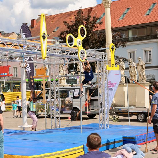 Eine Gruppe von Menschen spielt auf einem Hindernisparcours. Eine Person hängt an einem Seil, während andere zuschauen. Im Hintergrund befinden sich Statuen und Gebäude.
