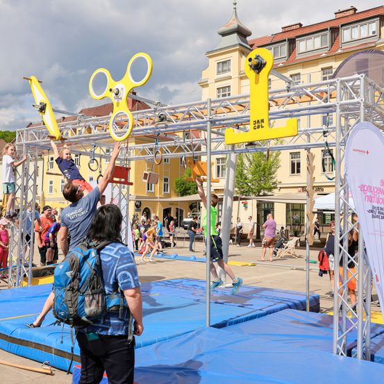 Eine Outdoor-Veranstaltung mit einer großen Hindernisparcours aus gelben Geräten. Teilnehmer klettern und schwingen an verschiedenen Elementen, während Zuschauer von den Seiten zusehen. Ein gelbes Gebäude und ein bewölkter Himmel bilden die Kulisse.