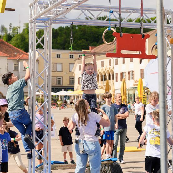 Eine Gruppe von Kindern und Erwachsenen spielt auf einem Metall-Hindernisparcours. Ein Mädchen hängt an einer Stange, und andere klettern auf Strukturen. Sie befinden sich in einem Außenbereich mit Gebäuden und Bäumen im Hintergrund.