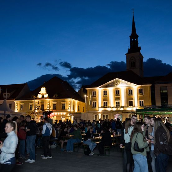Viele Menschen versammeln sich nachts vor einem beleuchteten Gebäude mit Turm, möglicherweise für eine Veranstaltung.