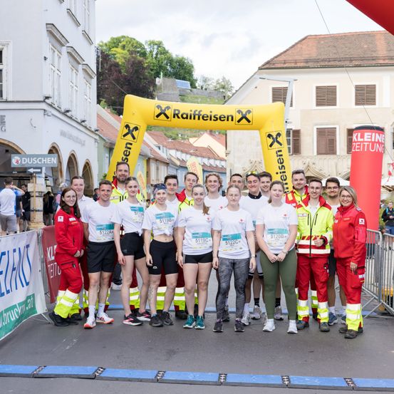 Eine Gruppe von Menschen, gekleidet in weißen T-Shirts und schwarzen Shorts, posiert für ein Foto vor einem gelben Raiffeisen-Banner. Sie sind wahrscheinlich Teilnehmer eines Marathons.