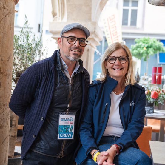 Ein Mann und eine Frau stehen und lächeln für ein Foto. Der Mann trägt einen Hut und eine Brille, und die Frau trägt ein weißes Hemd und Jeans. Sie befinden sich in einem Café mit Pflanzen und einem Tisch dahinter.