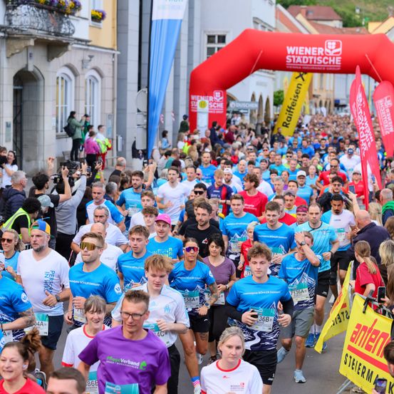 Eine große Menschenmenge läuft in einem Marathon. Die meisten tragen blaue Shirts. Im Hintergrund steht ein roter Bogen mit der Aufschrift 'Wiener Stadtische'. Zuschauer beobachten von der Seite.