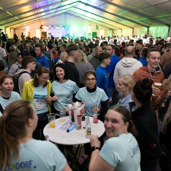 Eine große Menschenmenge versammelt sich in einem Zelt, viele tragen hellblaue Shirts mit Nummern und lächeln. Eine Frau hält ein Bier, und andere unterhalten sich um einen Tisch mit verschiedenen Getränken.