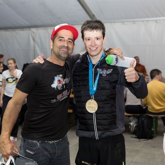 Zwei Männer stehen eng beieinander und lächeln für ein Foto. Einer trägt eine Medaille und hält eine Flasche, der andere einen roten Hut. Hinter ihnen sitzen und stehen Menschen, ein weißes Zelt im Hintergrund.