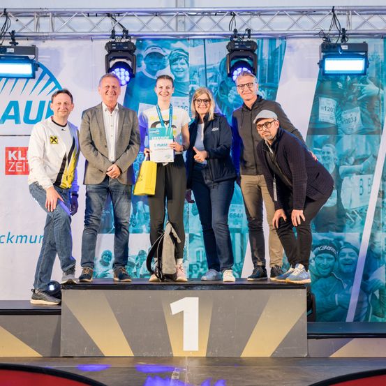 Fünf Personen stehen auf einem Podium, wobei eine Frau in der Mitte ein Zertifikat hält. Im Hintergrund ist ein großes Bild von Menschen beim Laufen zu sehen.