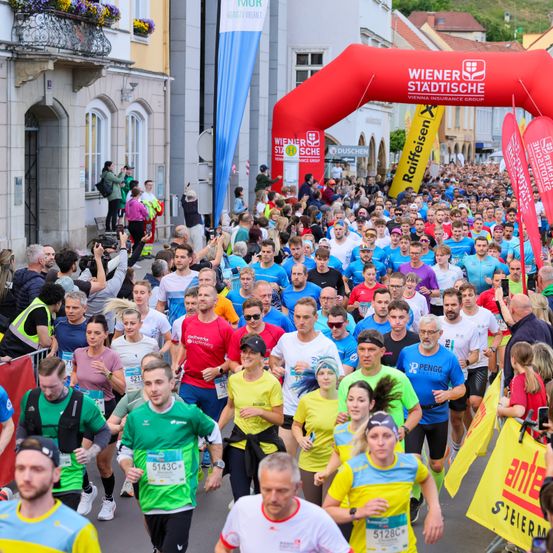 Eine große Menge von Läufern in verschiedenen farbigen Trikots und Nummern nimmt an einem Marathon teil und läuft durch ein Bannerbogen. Zuschauer jubeln ihnen zu.