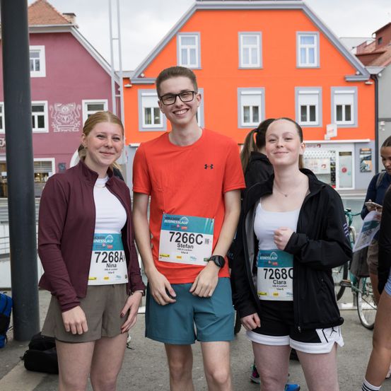 Drei Personen stehen vor einem leuchtend orangefarbenen Gebäude. Der Mann in der Mitte trägt ein oranges T-Shirt und eine Brille. Die Frauen auf beiden Seiten tragen burgunderfarbene und schwarze Jacken und haben sichtbare Startnummern.