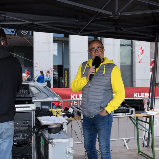 Ein Mann in einem gelben und grauen Jacken steht unter einem Zelt und spricht in ein Mikrofon. Hinter ihm ist eine Person in Schwarz und ein Auto zu sehen. Ausrüstung und Banner sind um ihn herum aufgestellt.