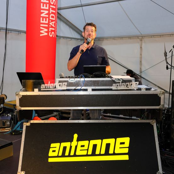 Ein Mann steht vor einem Mikrofon auf der Bühne, mit einem Laptop und einem Schild mit der Aufschrift 'Antenne' vor ihm. Er ist von Musikequipment und einem roten Banner umgeben.