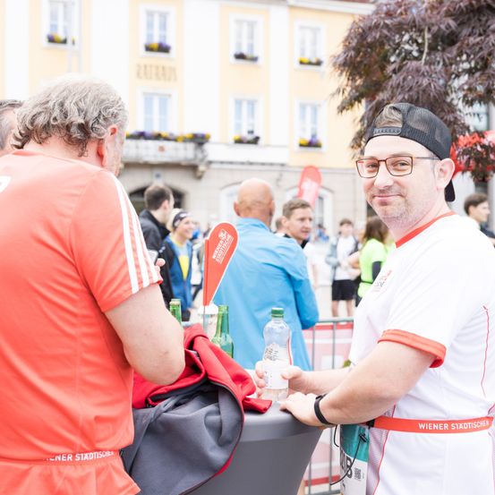 Ein Mann in einem weißen Shirt mit dem Wort 'Wiener' darauf erhält eine Wasserflasche von einem anderen Mann. Sie stehen vor einem Tisch mit Jacken darauf.
