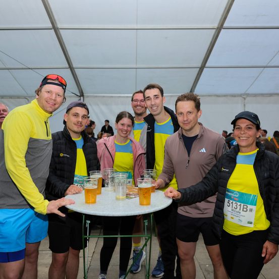 Eine Gruppe von Marathonläufern, gekleidet in Gelb und Grau, stehen um einen Tisch mit Gläsern Bier. Sie lächeln und posieren für ein Foto. Im Hintergrund scheinen Leute zu laufen.