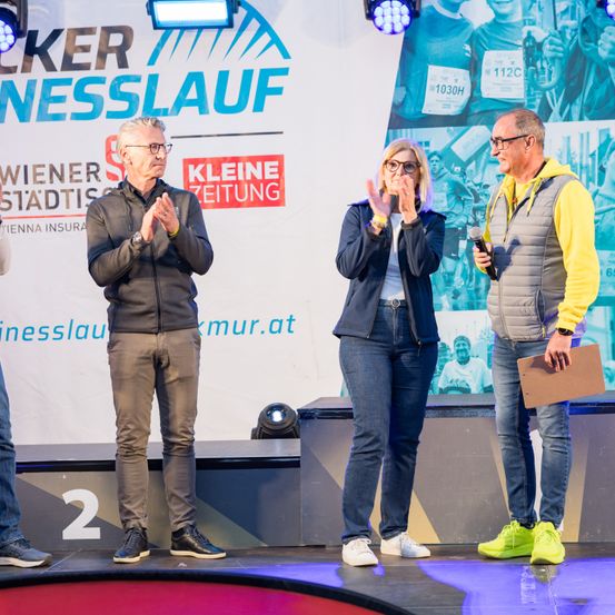 Drei Personen auf der Bühne mit einem großen Banner im Hintergrund. Der Mann auf der linken Seite klatscht, die Frau in der Mitte bedeckt ihren Mund und der Mann auf der rechten Seite hält ein Mikrofon.