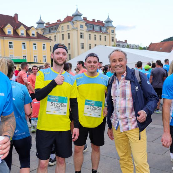 Eine Gruppe von Menschen versammelt sich im Freien, wahrscheinlich für einen Marathon. Zwei Männer tragen passende gelbe und blaue Trikots mit Startnummern. Ein älterer Mann in einem blauen Mantel und gelber Hose lächelt für die Kamera.