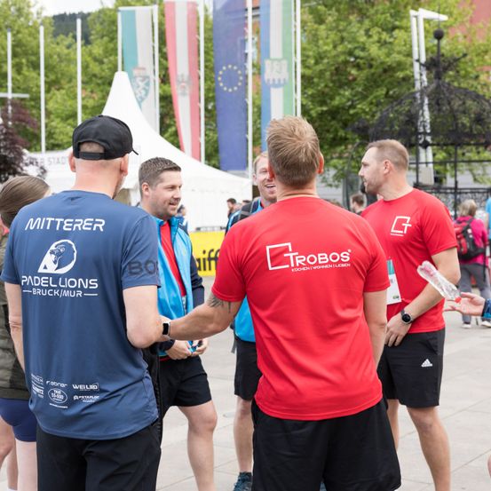 Eine Gruppe von Männern in verschiedenfarbigen T-Shirts unterhält sich im Freien. Ein Mann schüttelt einem anderen die Hand. Es gibt Leute, die herumlaufen, Fahnen und ein Zelt im Hintergrund.