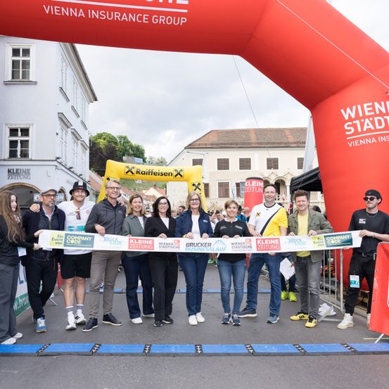 Menschen posieren für ein Foto vor einem roten Bogen mit einem Banner bei einer Veranstaltung. Dahinter sind Gebäude mit Bäumen und einem bewölkten Himmel.