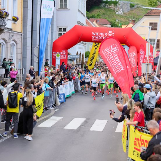 Ein Marathon findet in einer Stadt statt. Läufer laufen durch einen Bogen. Zuschauer säumen die Straße, einige machen Fotos. Fahnen und Banner sind sichtbar, Gebäude im Hintergrund.