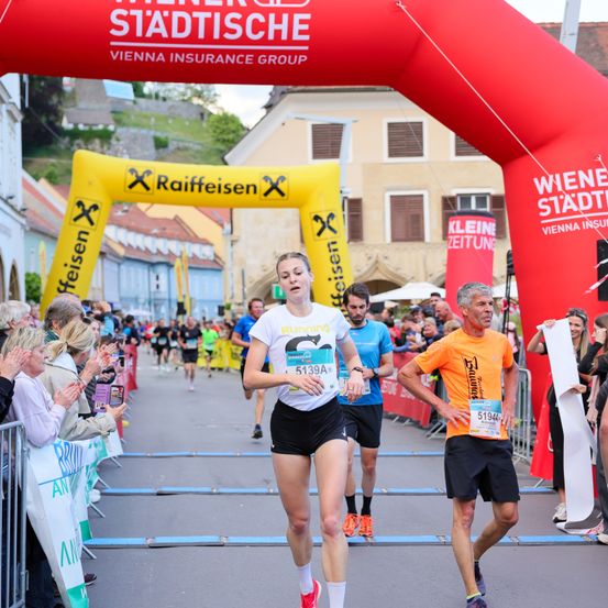 Läufer nehmen an einem Marathon teil. Eine Frau in einem weißen Trikot mit einer Nummer ist vor einem Mann in einem orangefarbenen Trikot. Die Menge jubelt ihnen zu.