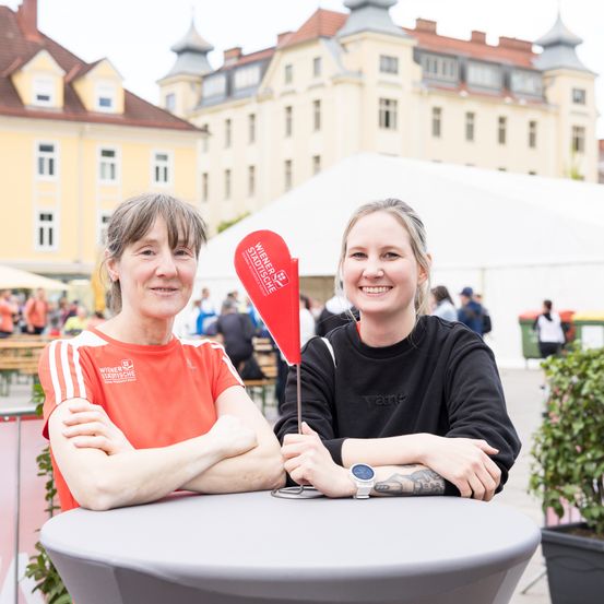 Zwei Frauen sitzen an einem Tisch und posieren lächelnd für ein Foto. Die Frau auf der linken Seite trägt ein orangefarbenes Shirt mit weißen Streifen, und die Frau auf der rechten Seite hat eine Uhr und Tattoos. Hinter ihnen ist ein Gebäude mit vielen Fenstern zu sehen, zusammen mit Menschen und einem Zelt.