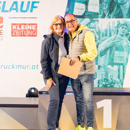 Zwei Personen auf der Bühne, die eine Mappe halten und für ein Foto lächeln, mit einem Hintergrund eines Rennensposters.