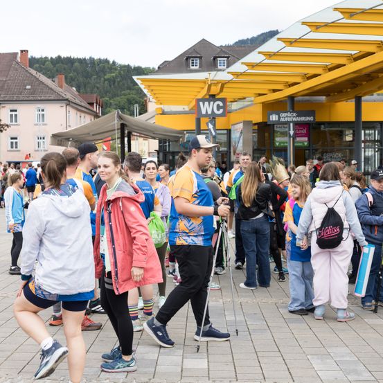 Eine Menschenmenge in Sportbekleidung, wahrscheinlich Marathon-Teilnehmer, versammelt sich vor einem Gebäude mit gelben Vordächern. Einige halten Gehstöcke. Im Hintergrund sind Gebäude und Berge zu sehen.