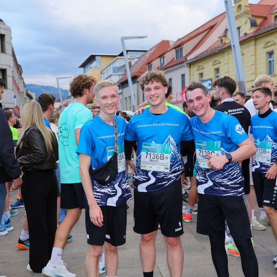 Eine Gruppe von Marathonläufern posiert für ein Foto. Die Männer in blauen Shirts tragen Nummern, die ihre Startnummern anzeigen. Sie stehen unter einer größeren Gruppe von Läufern, die alle passende blaue Shirts tragen. Der Hintergrund zeigt Gebäude und Berge.