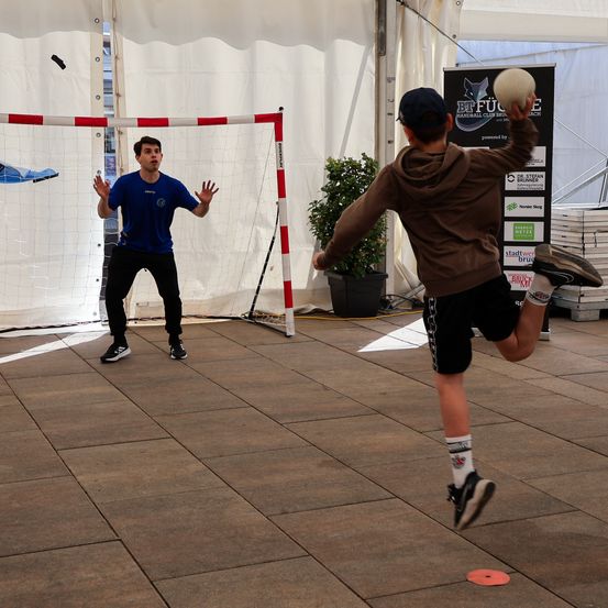 Zwei Männer spielen Futsal auf einem Platz mit einem Tor. Ein Mann hält einen Ball bereit zum Kicken. Ein Topf mit Pflanze steht in der Nähe.