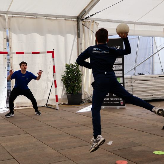 Zwei Männer spielen mit einem Ball in einem Zelt. Ein Mann bereitet sich auf den Wurf vor, während der andere blockt. Dahinter steht ein weißes Netz und ein Topf mit einer Pflanze.
