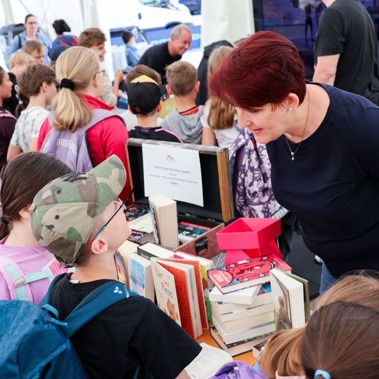 Eine Frau mit roten Haaren spricht mit einem jungen Jungen auf einem Büchermarkt. Andere Kinder und Erwachsene sind um sie herum und stöbern durch die ausgestellten Bücher.