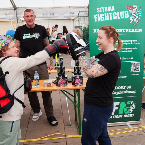 Eine Frau boxt mit einem Kind vor einem Banner, das für den Styrian Fightclub wirbt. Ein Mann steht hinter ihnen und trägt ein Shirt mit dem Logo des gleichen Clubs.