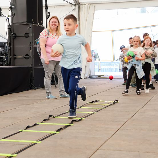 Ein Junge läuft mit einem Ball in der Hand durch ein Hindernisparcours, während eine Frau zusieht. Andere Kinder warten hinter ihm mit farbigen Bällen. Ausrüstung steht auf der Bühne, und es gibt ein Banner an der Wand.