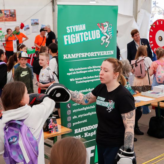 Eine Frau mit Tätowierungen unterrichtet ein junges Mädchen im Boxen bei einer Veranstaltung des Syrian Fight Club. Eine Menschenmenge beobachtet, und ein grüner Banner mit Clubinformationen ist sichtbar.