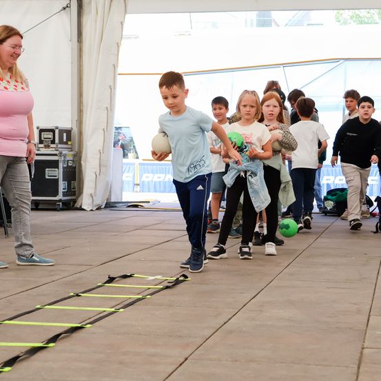 Eine Gruppe von Kindern läuft durch einen Hindernisparcours und hält verschiedene Bälle in der Hand. Eine Frau steht daneben und beobachtet sie. Die Szene spielt sich unter einem Zelt mit weißem Hintergrund ab.