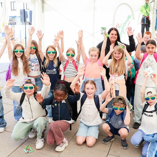 Eine Gruppe von Kindern und einer erwachsenen Frau, die Sonnenbrillen tragen, posiert für ein Foto mit erhobenen Händen. Sie befinden sich in einem Raum mit einem weißen Zelt und einem großen weißen Bogen. Einige von ihnen tragen Rucksäcke und Turnschuhe. Dahinter klettert eine Person in einem grünen Shirt an einem Seil.