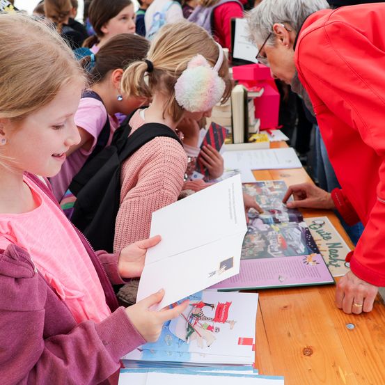 Ein kleines Mädchen hält ein Buch, während eine ältere Frau es betrachtet. Andere Kinder sind am Tisch und schauen sich andere Bücher an. Die Frau trägt eine rote Jacke.