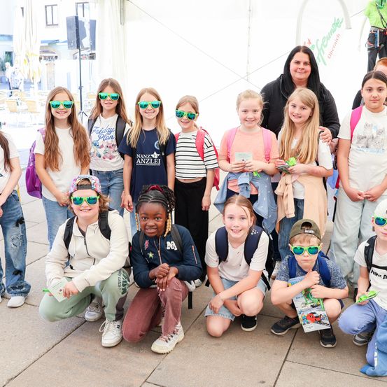 Eine Gruppe junger Mädchen und Jungen mit Sonnenbrillen und Rucksäcken, wahrscheinlich auf einer Klassenfahrt, lächeln für die Kamera. Sie stehen auf einem Betonboden.