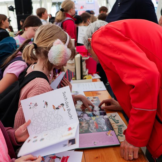 Eine Gruppe von Menschen, einschließlich Kindern, untersucht Bücher an einem Tisch. Ein Mädchen mit rosa Ohrenschützern hält ein Buch. Ein Mann in einem roten Mantel beugt sich über den Tisch.
