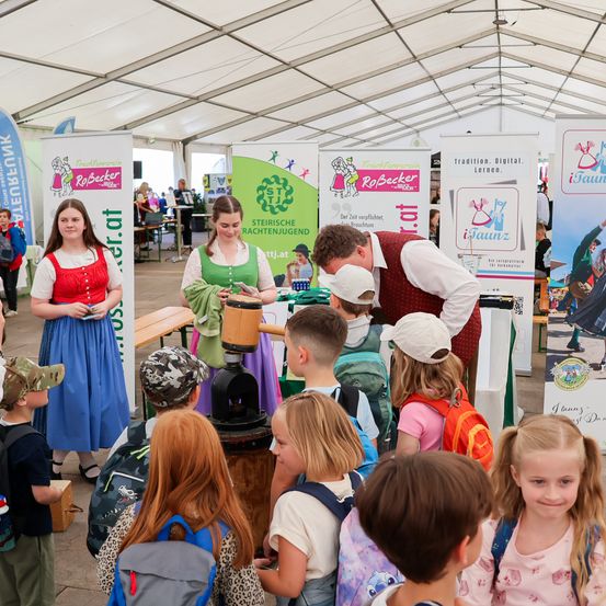 Eine Gruppe von Kindern und Erwachsenen steht um eine Holzpresse in einem Zelt herum. Zwei Frauen in traditioneller Kleidung stehen in der Nähe. Einige Kinder tragen Rucksäcke.