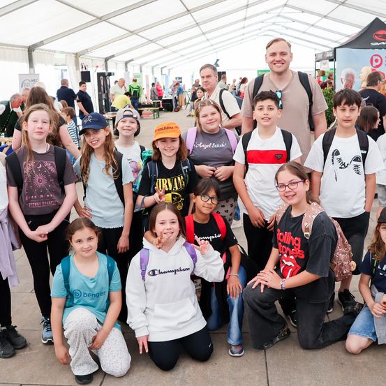 Eine Gruppe von Kindern und Jugendlichen posiert für ein Foto unter einem Zelt mit Menschen und Ständen im Hintergrund.