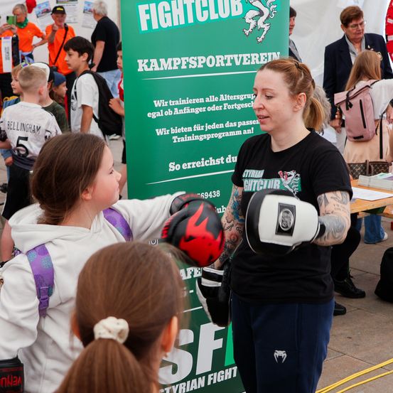 Eine Frau mit Tätowierungen und Boxhandschuhen spricht mit einem jungen Mädchen. Ein Schild hinter ihnen sagt 'FIGHTCLUB'. Im Hintergrund sind Kinder und Erwachsene zu sehen.
