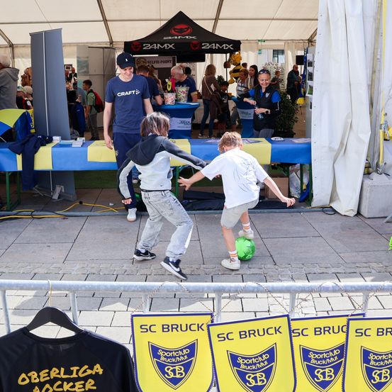 Zwei Kinder spielen mit einem Fußball auf einem gepflasterten Bereich. Eine Person in einem blauen T-Shirt steht hinter ihnen. Ein Tisch mit einem blauen Tuch und gelben Akzenten steht vor ihnen. Es gibt Banner und ein Zelt mit dem Text 24MX im Hintergrund.