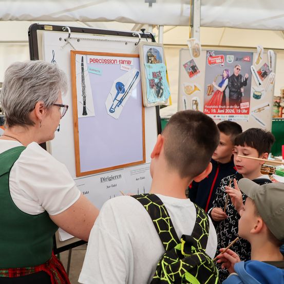 Eine ältere Frau mit Brille zeigt einer Gruppe von Jungen ein Poster mit Musikinstrumenten. Das Poster hat das Wort 'Perkussionsfamilie' und die Jungen halten Chopsticks.