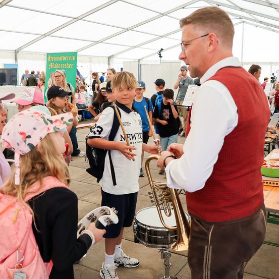 Ein Mann in einem roten Vest demonstriert ein Kind eine Trompete, ein Kind hält ein Tamburin. Die Kinder sind in einem Zelt im Freien.