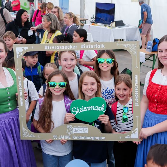 Kinder und Erwachsene posieren vor einem Fotorahmen bei einer Outdoor-Veranstaltung. Der Rahmen hat ein grünes Herz und den Text Steiermark. Die Kinder tragen Sonnenbrillen.
