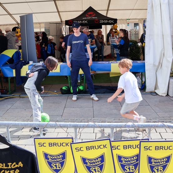 Ein Mann steht in einem Zelt, während Kinder auf einer Plattform mit einem Ball spielen. Die Plattform hat Banner für SC Bruck. Menschen stehen herum, einige in der Nähe eines Tisches.