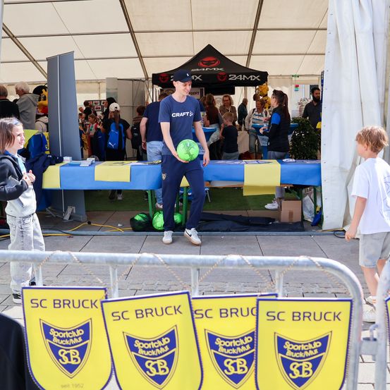 Ein Mann in einem blauen T-Shirt steht unter einem Zelt und hält einen grünen Fußball, während eine Gruppe von Menschen im Hintergrund beobachtet. Viele Menschen sind anwesend, einige in Sportbekleidung. SC Bruck-Banner sind an einem Zaun angebracht.