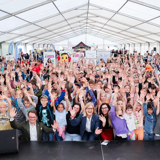 Eine Menschenmenge, bestehend aus Kindern und Erwachsenen, versammelt sich unter einem weißen Zelt für ein Gruppenfoto. Sie heben ihre Hände, einige tragen Brillen, Mützen und Rucksäcke. Ein Lautsprecher und ein Banner sind links.