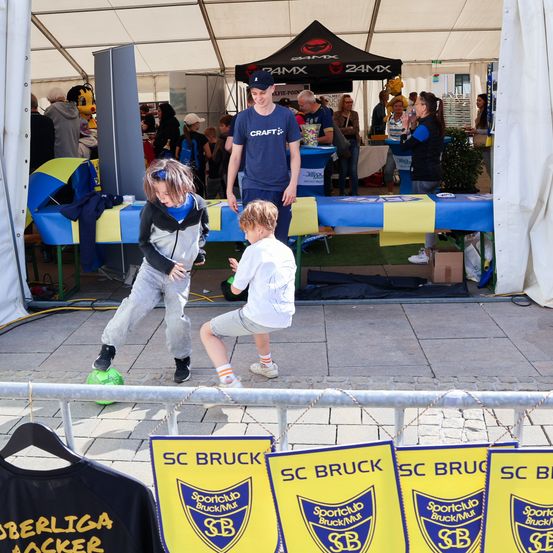 Zwei Kinder spielen Fußball mit einem Ball auf einer grünen Oberfläche. Ein Mann beobachtet sie von hinten. Es gibt mehrere Banner und einen Tisch mit Gegenständen darauf. Im Hintergrund stehen Menschen in der Nähe eines Zeltes.