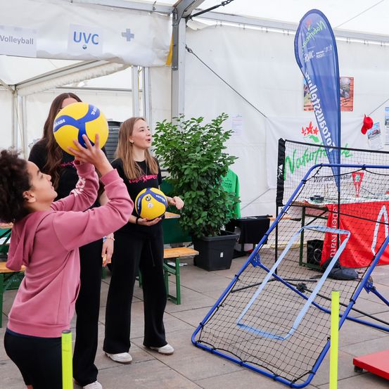 Drei Frauen spielen Volleyball in einem Zelt mit Netz und blauer Fahne. Das Mädchen in Pink hält einen Ball, während ein anderes Mädchen einen Ball in der Nähe einer Pflanze hält.
